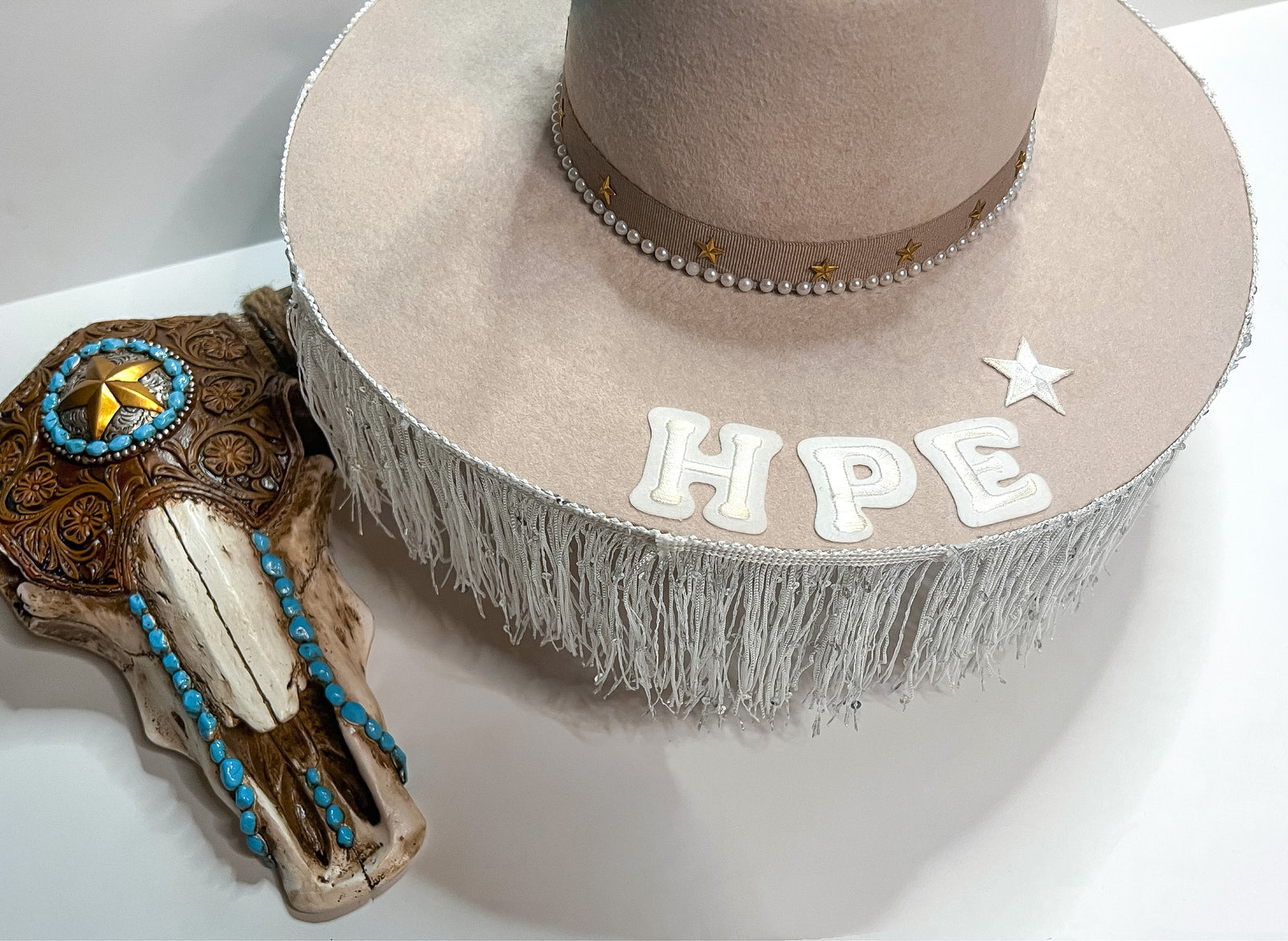 Pictured is a tan felt hat with white fringe on the brim, HPE in white felt letters, ine white star, pearls and gold stars around the crown of the hat.