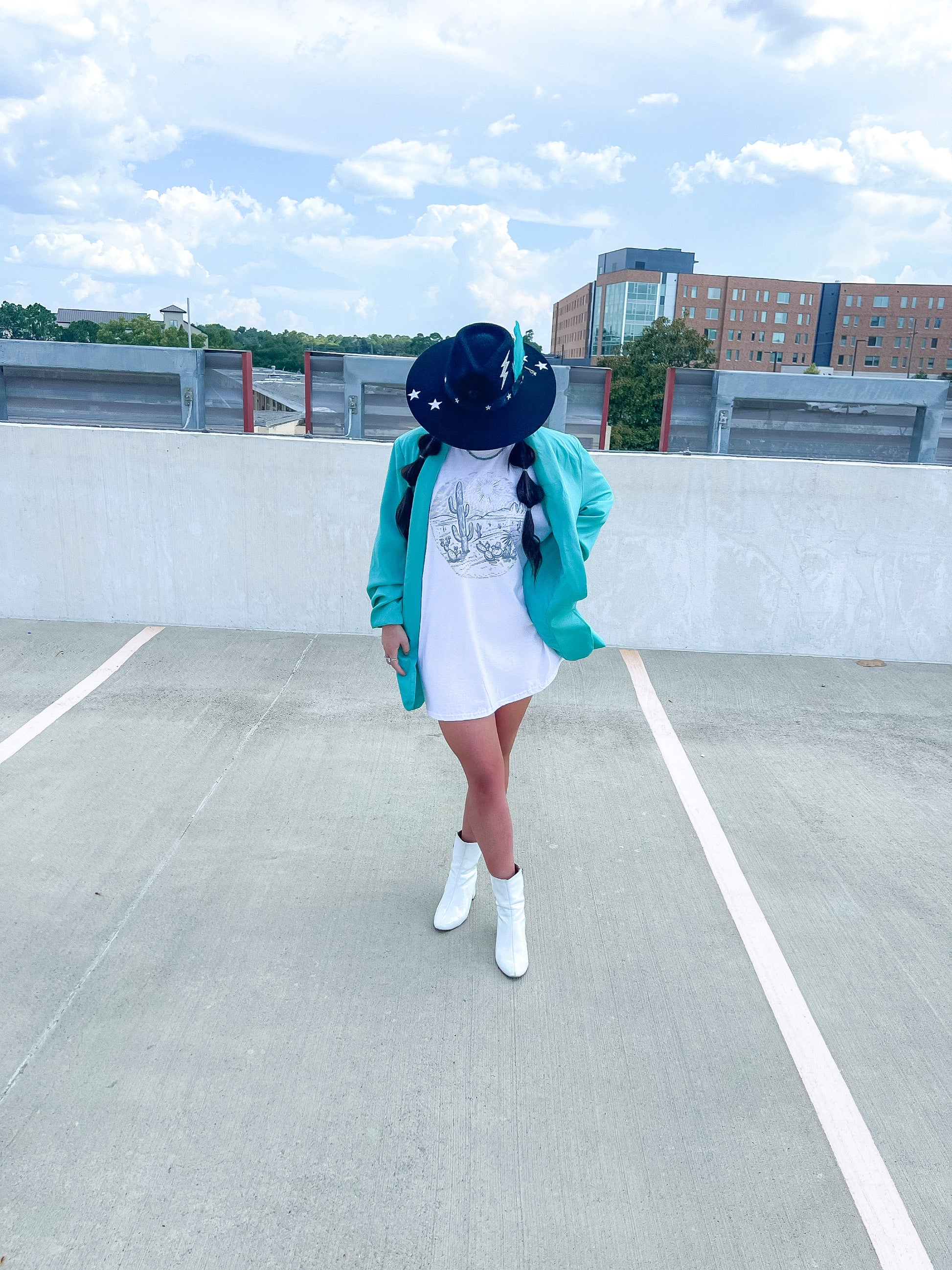 Pictured is a dark headed girl wearing a black hat, teal blazer, white graphic tee and white booties. She is pictured outside looking at her feet.